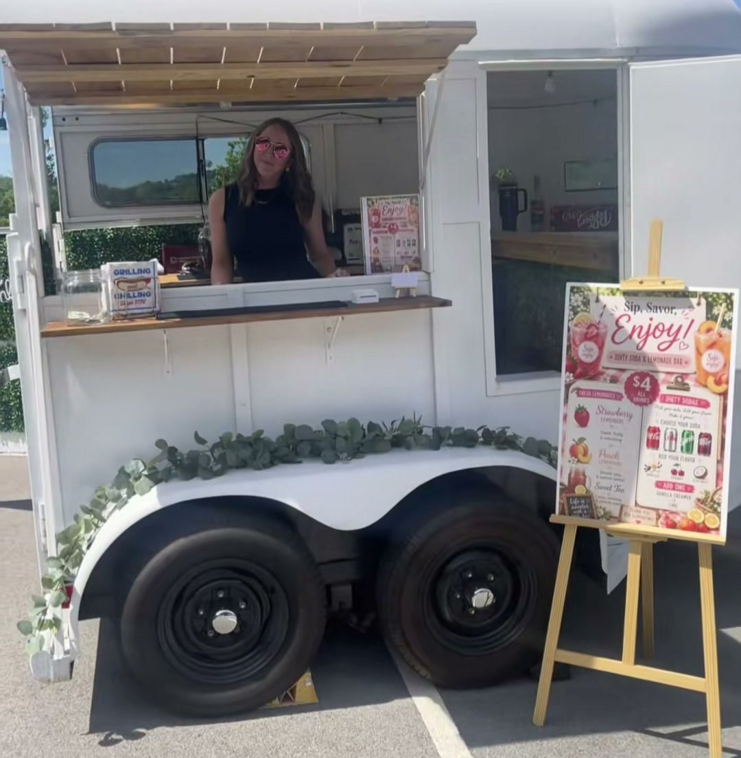 The trailer set up at an event with the awning open, eucalyptus along the fender and a printed menu on an easel
