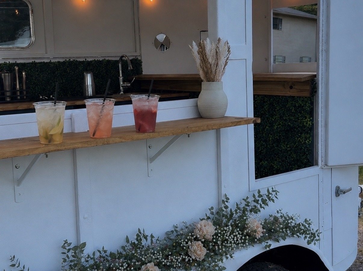 Three drinks on the trailer's wooden bar shelf under warm string lights